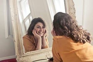 pexels-photo-774866-774866 A cheerful woman smiles at her reflection in a vintage-style mirror, exuding positivity and warmth.